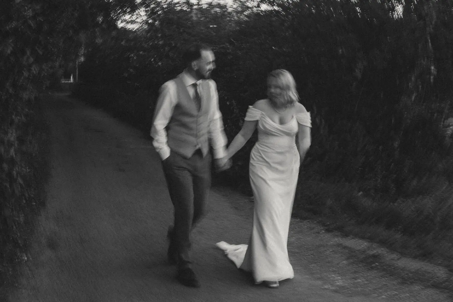 Bride and groom walking hand in hand along a tree-lined lane in a blurred black and white photograph