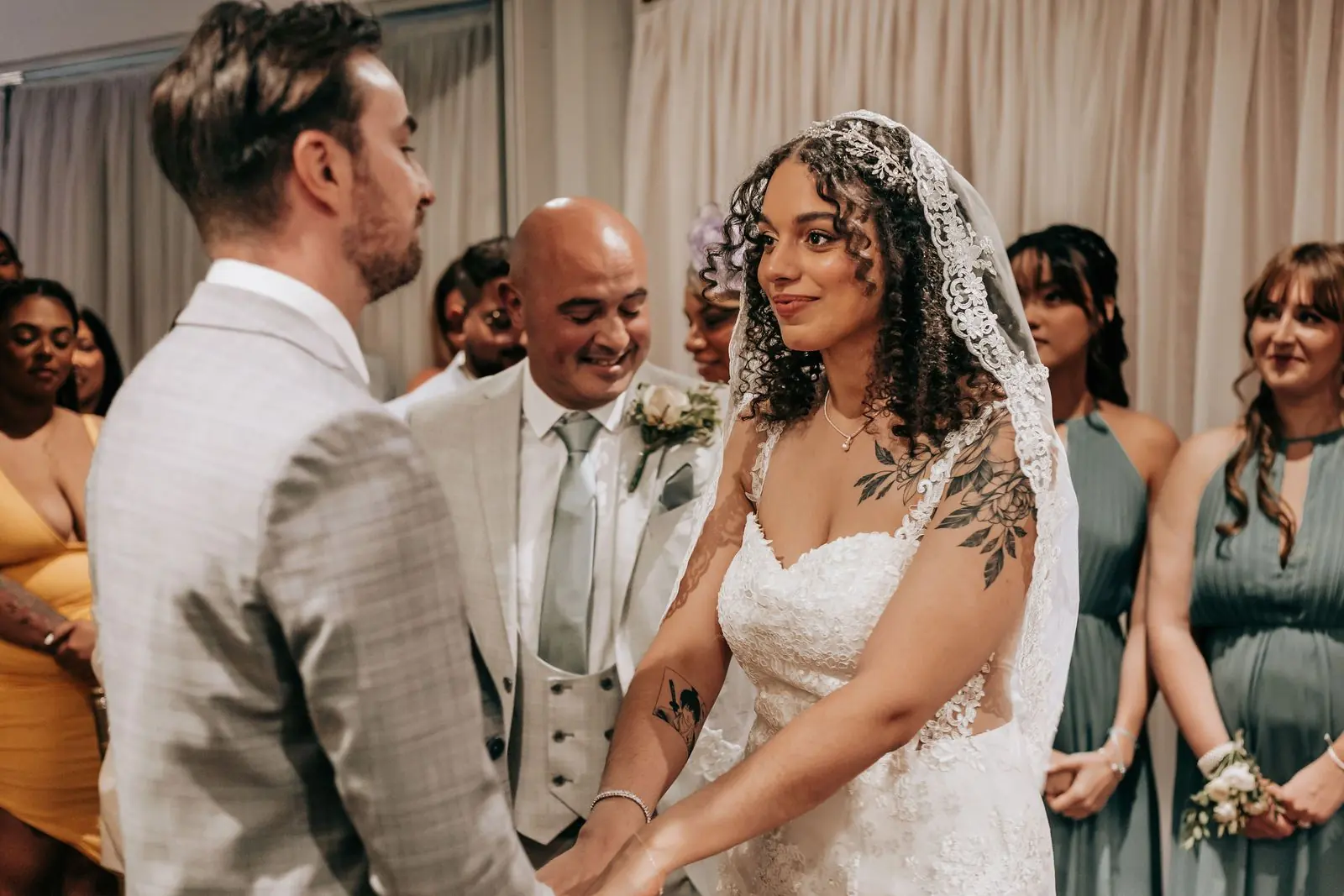 Bride in a lace veil during the ceremony as the groom looks on